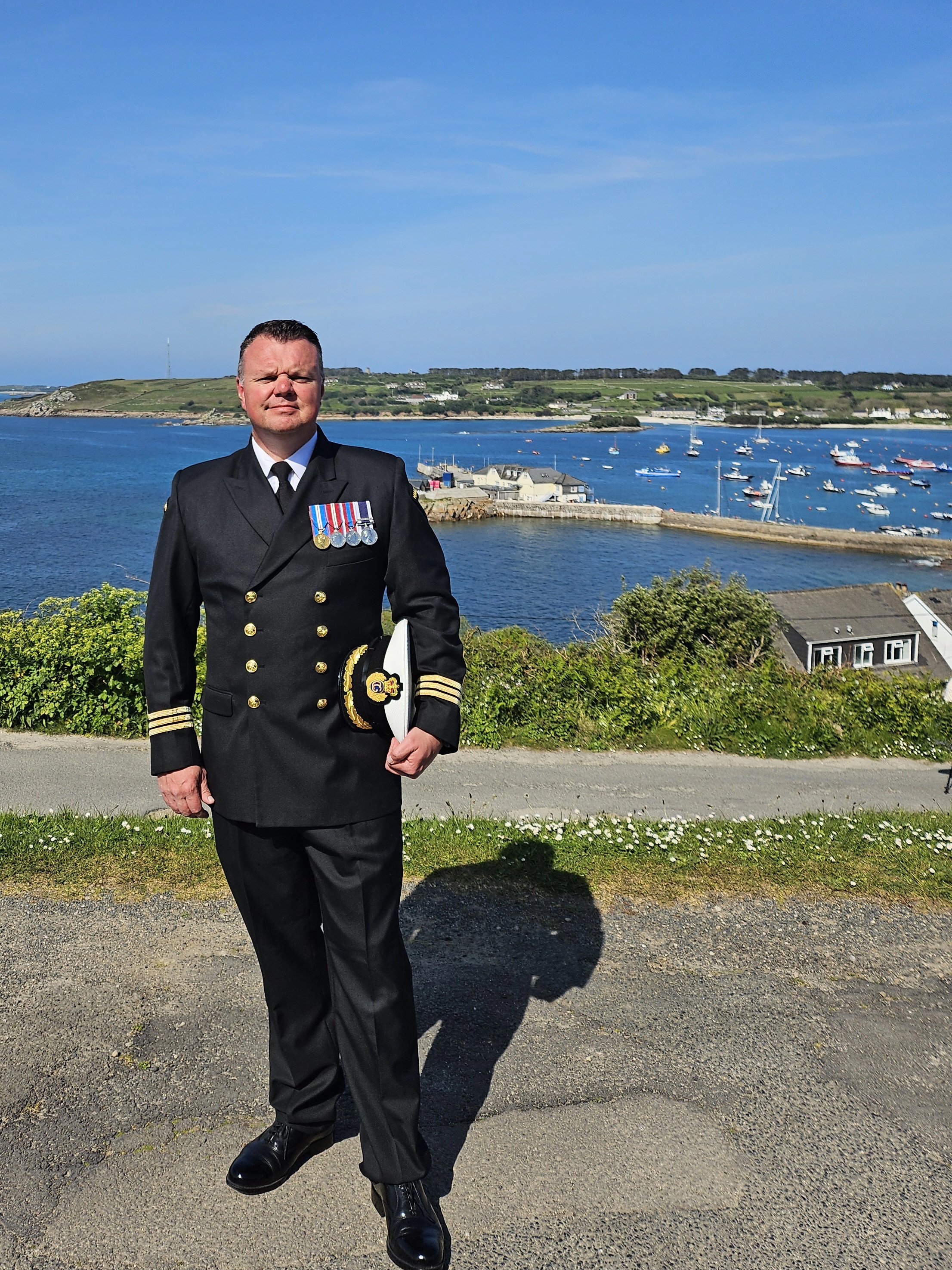 Jon Oliver stood in Royal Navy uniform at a coastal location on a sunny day