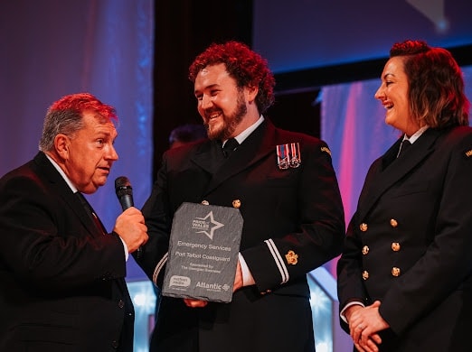 On the right, a male and a female coastguard rescue officer in formal attire stand receiving an award on a stage. On the right, a male dressed in a suit is holding a microphone