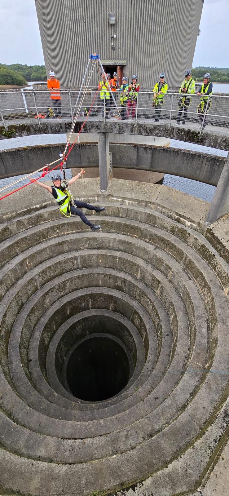 A woman wearing harness and helmet dangles from a climbing rope strung across a deep, concrete-lined hole
