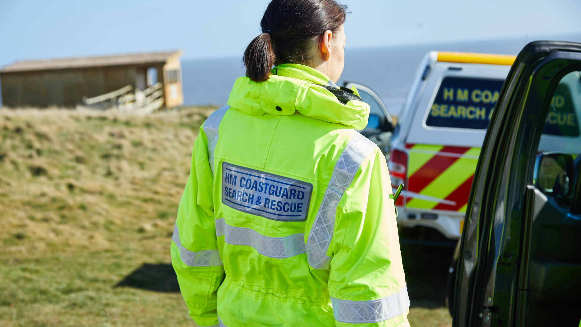 A female coastguard in high-viz jacket stands near a vehicle by a cliff