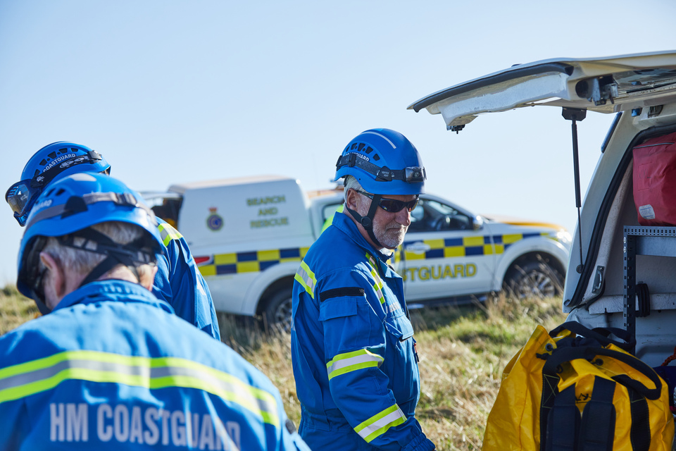 A coastguard in blue overalls stands by the back of a vehicle