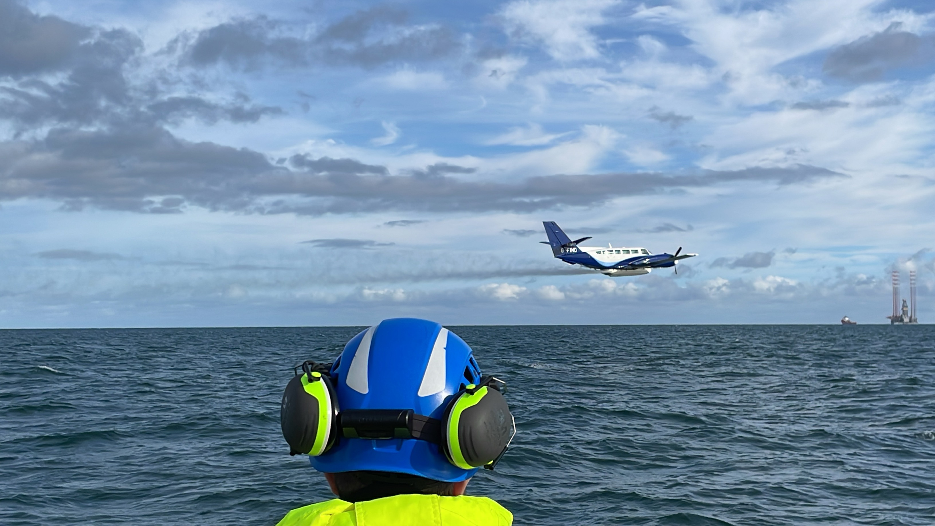 A helmeted man watches a plane fly past low over the sea surface