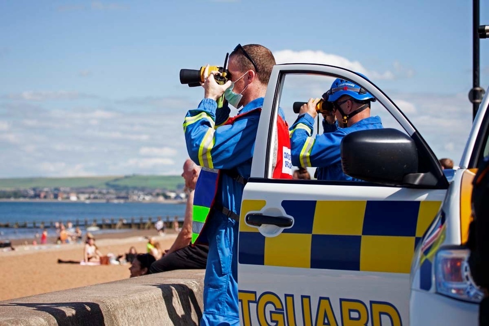 Two coastguards with binoculars look out across a beach