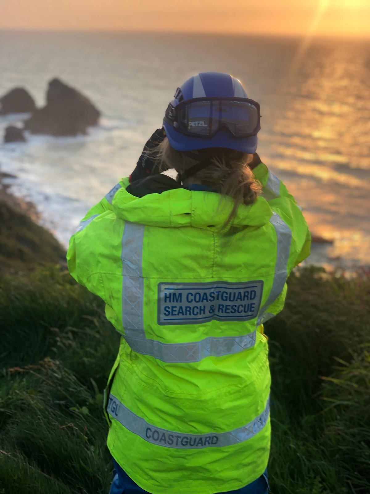 A Coastguard in hi-viz jacket looks out through binoculars from the cliff out to sea