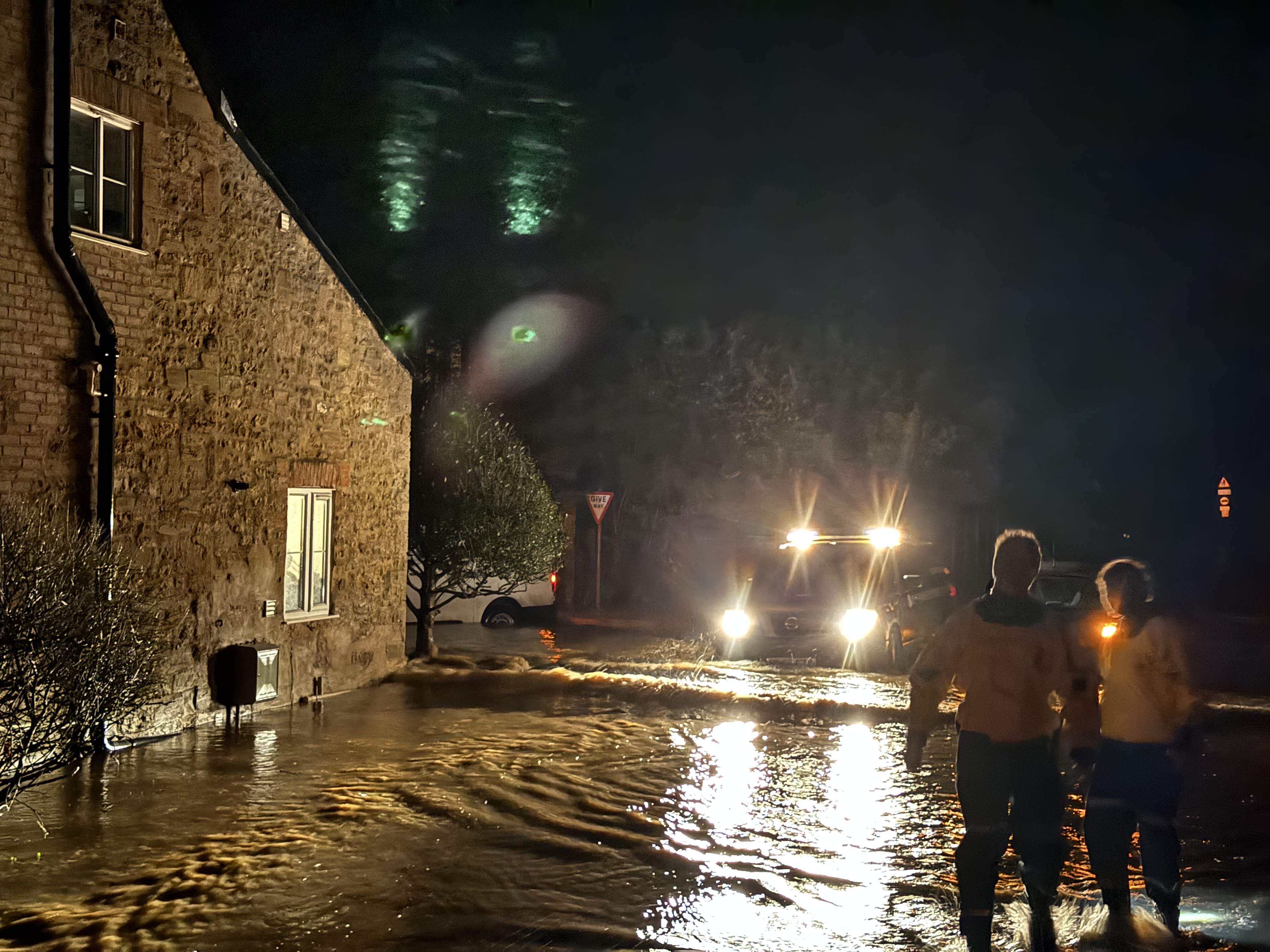 Two Coastguard Rescue Officers wading in a flooded street are silhouetted by vehicle lights