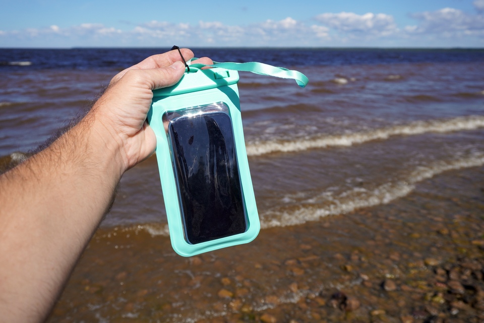 A hand holding a mobile phone in a green waterproof case while on a pebbly beach with sea waves in the background