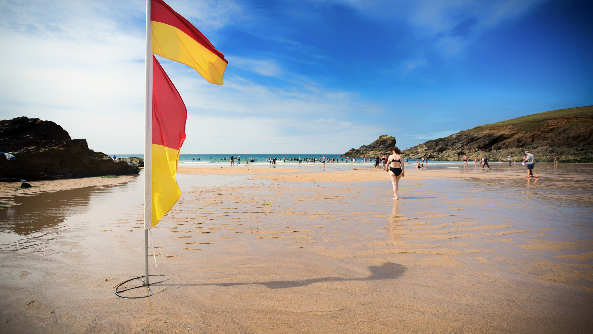 A red and yellow flag flaps at a beach as a woman walks towards the water