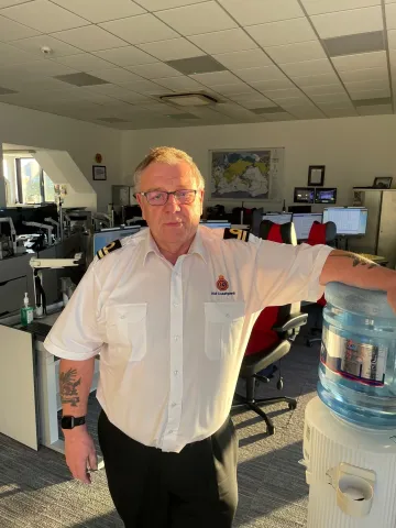 Unsmiling man in Coastguard uniform stood in front of camera in operations room.