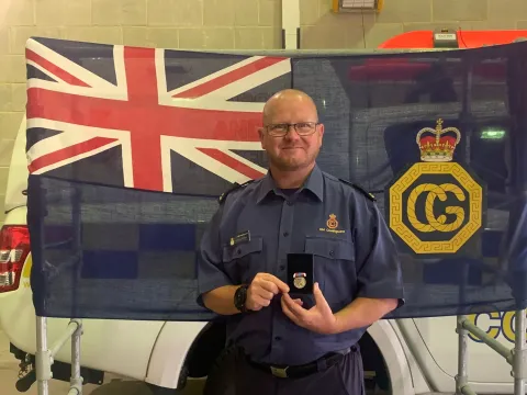 Coastguard Rescue Officer Kevin Moran in uniform stands in front of an HM Coastguard flag