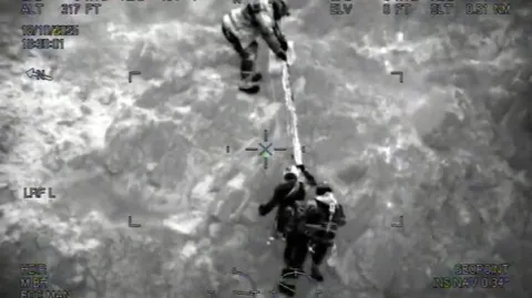 Night-vision image of a climber on a rope being helped up the cliff by two Coastguard Rescue Officers