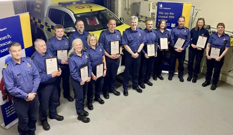 Smiling Coastguards in blue uniforms hold their awards with Assistant Chief Coastguard John Craig standing in the middle
