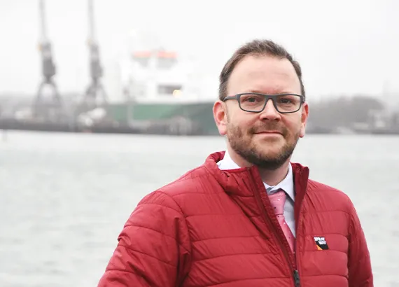 Portrait photo of Stephan. He is wearing a red jacket over a shirt and tie, stood in front of a body of water with docks in the distant background.