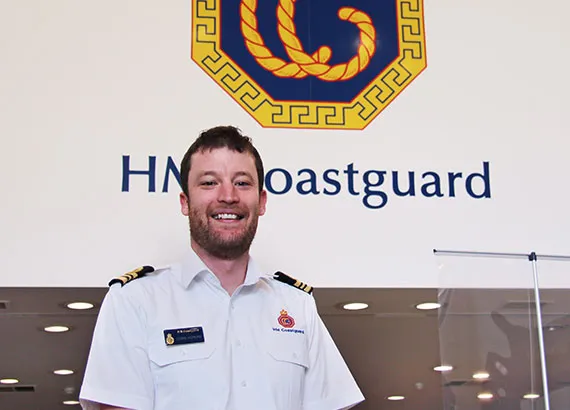 Chris Hopkins stood in the Joint Rescue Co-ordination Centre. In the background there is a large HM Coastguard logo on the wall.