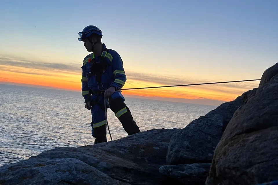 A coastguard roped to a cliff looks out to sea silhouetted by a sunset