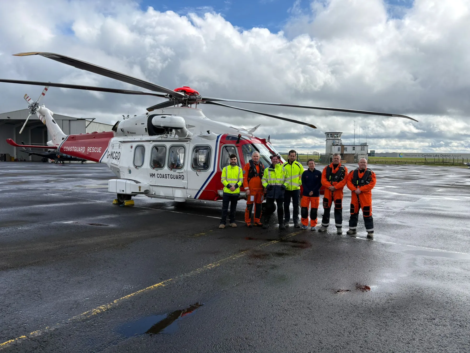 Members of the AW189 helicopter crew at Carlisle, some in orange overalls and others in high-visibility jackets, standing in front of a white and red helicopter. From left to right: Frank Napier (Engineer), Rich Taylor (Winch Operator), Michael Walker (Engineer), Daniel Perry (Engineer), Cathy Freeman-Brown (Winch-Paramedic), Simon White (Captain), Simon Price (Co-Pilot).