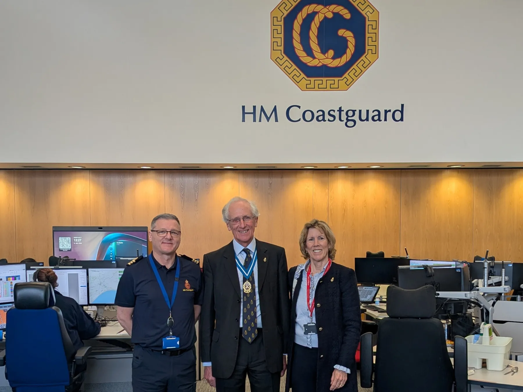 In the JRCC operations room with an HM Coastguard logo behind them stand Matt West, William Maltby and Sarah Maltby