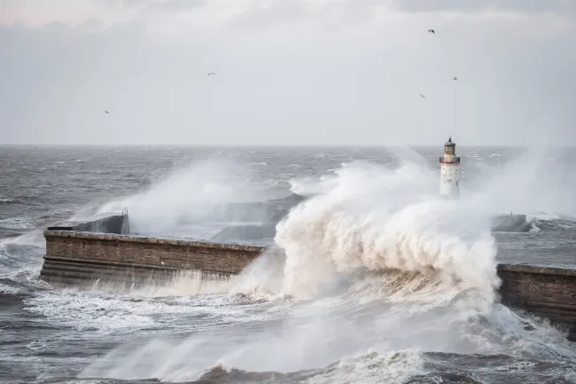 Storm waves crash over harbour wall and lighthouse in stormy seas