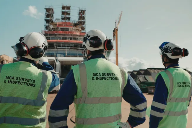 Three surveyors in high-vis and hard hats look up at the bridge of a vessel