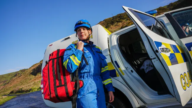 A female coastguard carrying a red bag stands near a vehicle