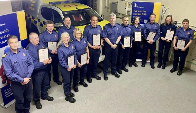 Smiling Coastguards in blue uniforms hold their awards with Assistant Chief Coastguard John Craig standing in the middle
